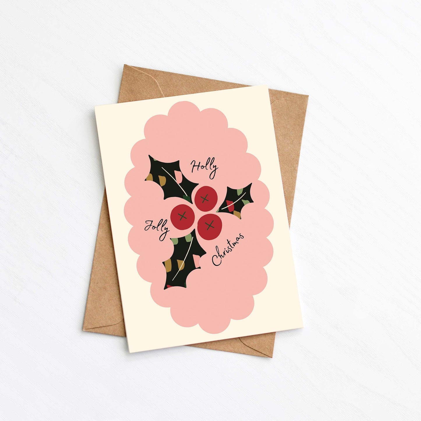 Flat lay of a modern Christmas card featuring a pink scalloped frame, red holly berries, and dark green holly leaves with the text "Holly Jolly Christmas," placed on a kraft envelope.