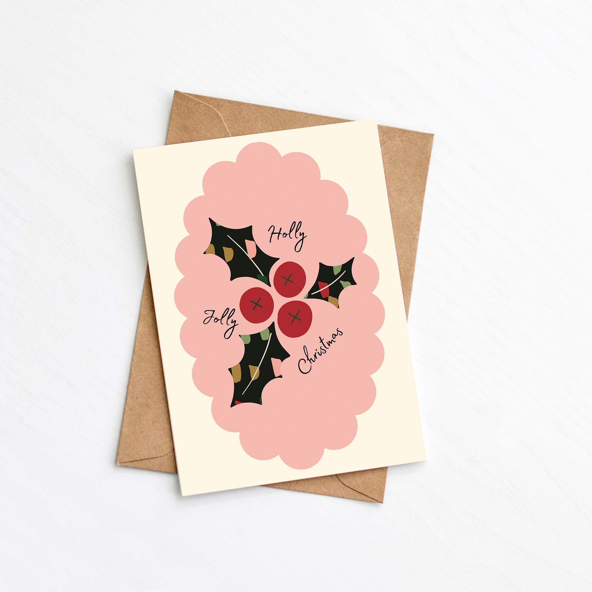 Flat lay of a modern Christmas card featuring a pink scalloped frame, red holly berries, and dark green holly leaves with the text "Holly Jolly Christmas," placed on a kraft envelope.