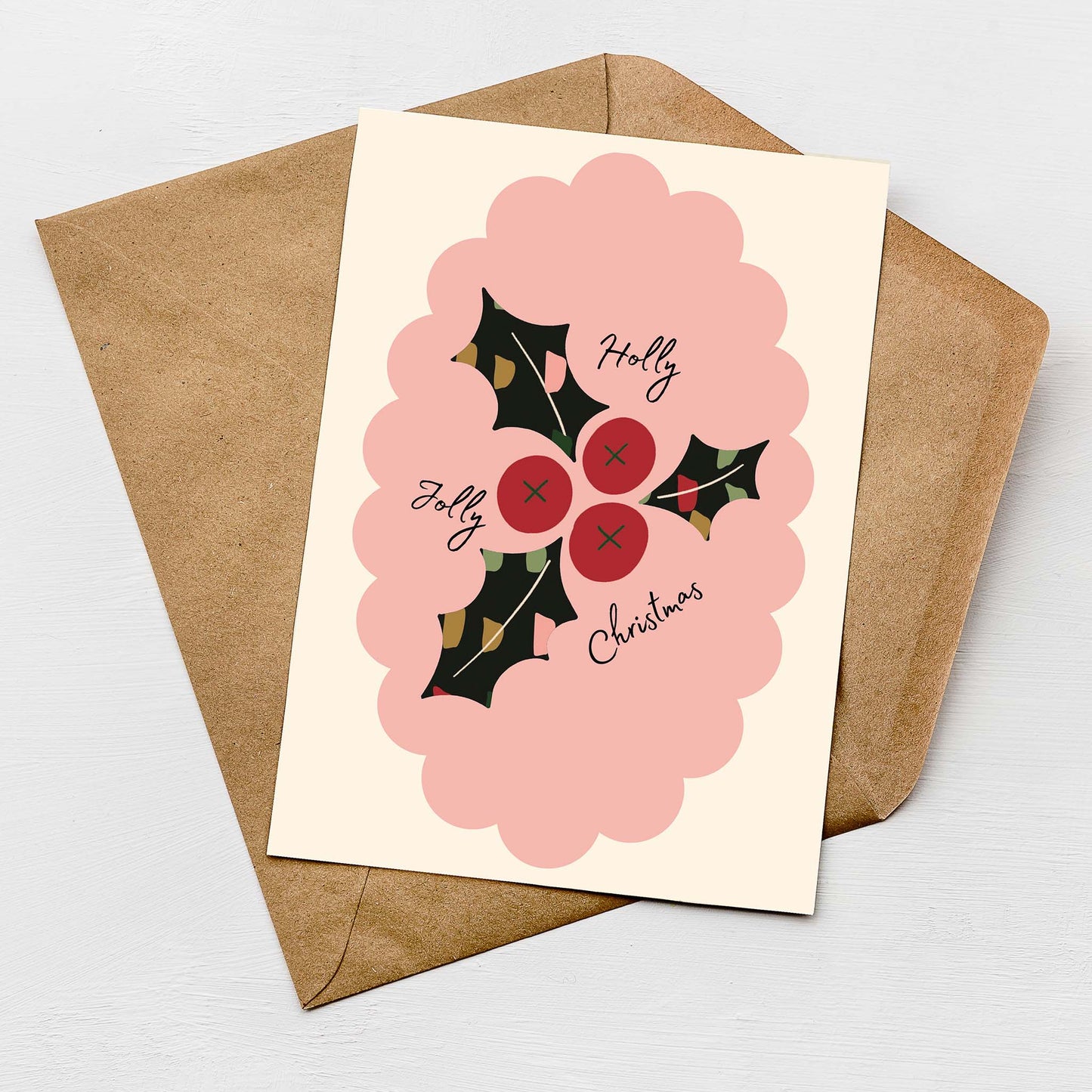 Flat lay of a modern Christmas card featuring a pink scalloped frame, red holly berries, and dark green holly leaves with the text "Holly Jolly Christmas," placed on a kraft envelope.