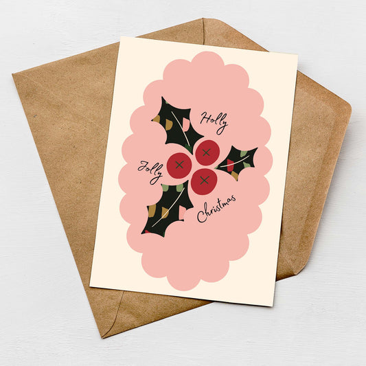 Flat lay of a modern Christmas card featuring a pink scalloped frame, red holly berries, and dark green holly leaves with the text "Holly Jolly Christmas," placed on a kraft envelope.