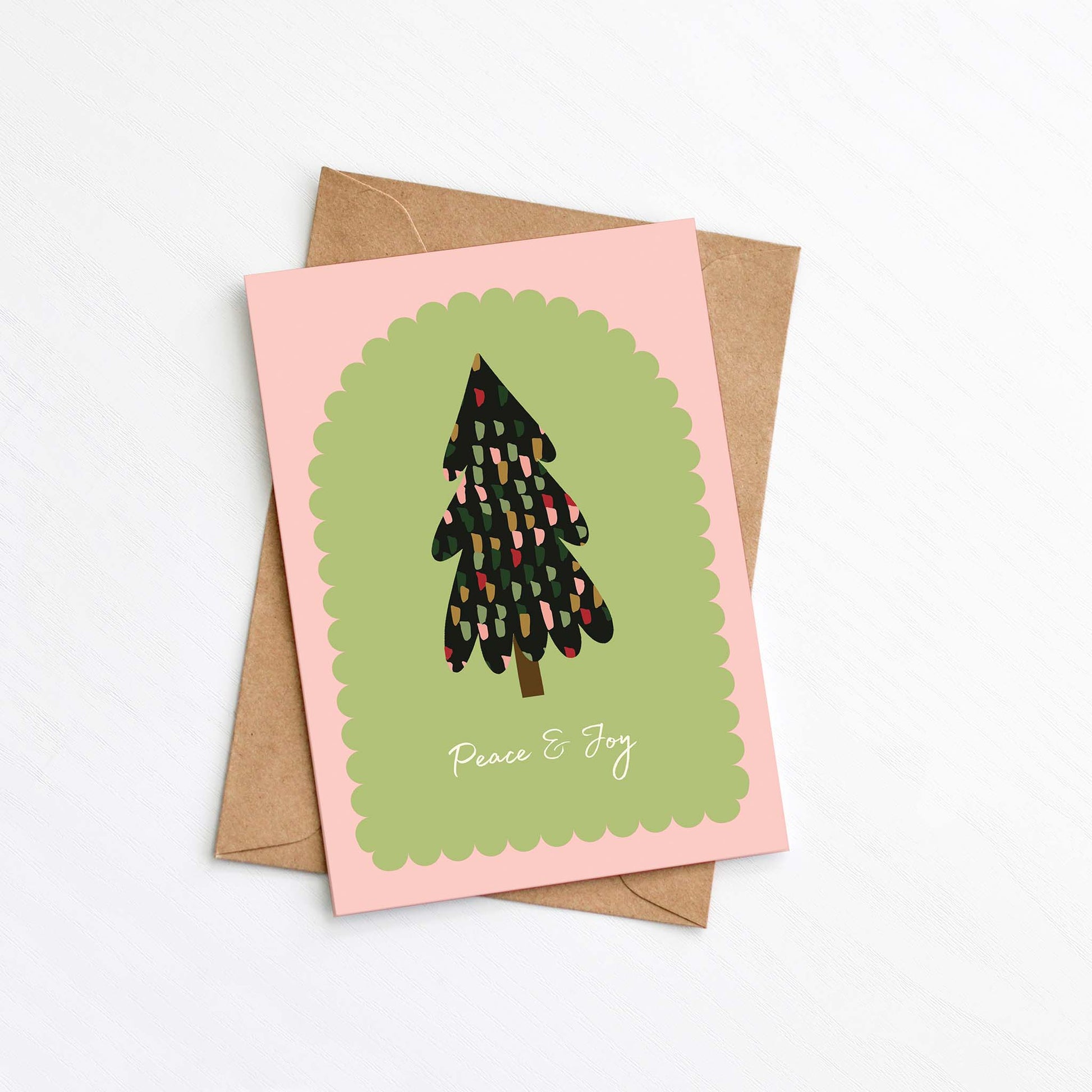 Peace & Joy Christmas card with illustrated tree and kraft envelope on pink and green background